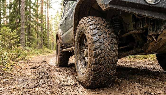 Close-up of a muddy all-terrain tire on a vehicle driving on a rugged dirt trail with forest in the background