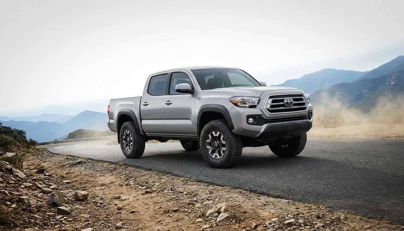 Silver pickup truck with all-terrain tires driving on a road next to a rocky dirt path in a mountainous landscape