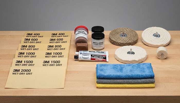 Range of polishing compounds wet-dry sandpapers in multiple grits buffing wheels and pads laid out on a workbench for aluminum wheel restoration
