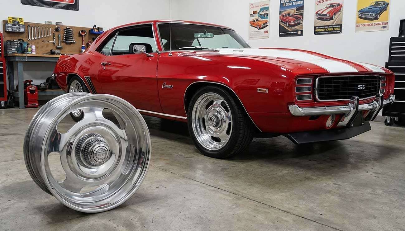 Silver five-spoke American Racing Rally wheel with chrome lip beside a red classic muscle car