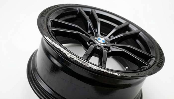 Gloss black wheel with visible curb rash exposing bare aluminum on the outer lip against a clean white studio backdrop