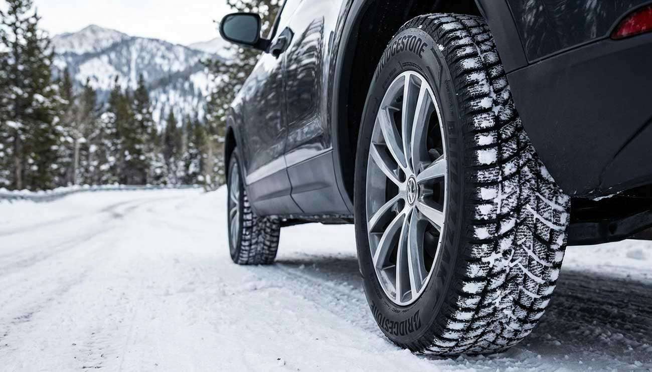 Close-up of a vehicle's Blizzak winter tire gripping a snowy road in a mountainous, forested winter landscape