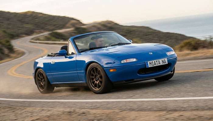 Blue NA Mazda Miata with aftermarket wheels driving on a road with the driver leaning out the window