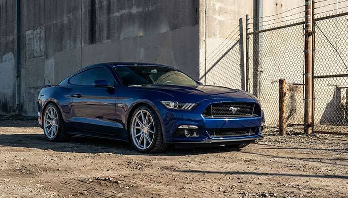 Blue S550 Ford Mustang with Velgen wheels parked near a concrete wall under a metal fence on a dirt surface