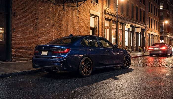 Blue BMW M340i with black staggered wheels parked in a nighttime lot near a brick building