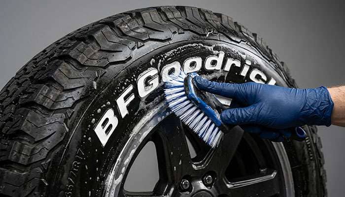 Hand using a stiff bristle brush to scrub raised white letters on a tire sidewall during detailed cleaning with soapy water
