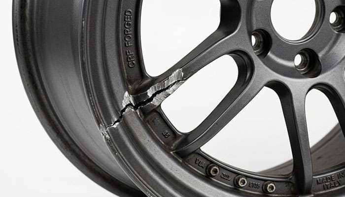 Macro close-up of a cracked aluminum wheel barrel showing a visible structural fracture line at the spoke-to-barrel junction against a clean white studio backdrop