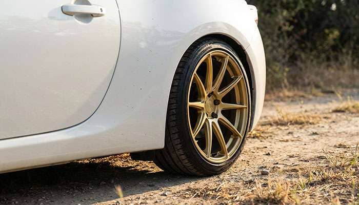 Close-up of a white car highlighting custom gold aftermarket wheels against a dirt ground background