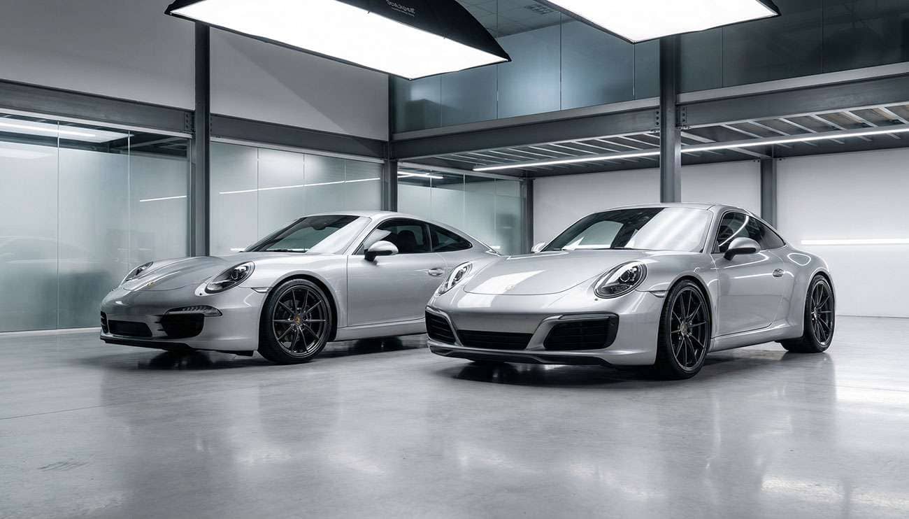 Two silver sports cars with custom offset wheels parked side by side in a garage with bright overhead lighting