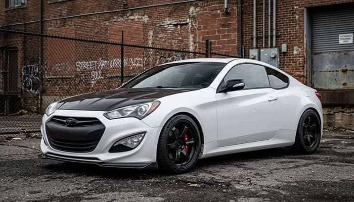 White Hyundai Genesis Coupe with black hood and aftermarket wheels parked in front of a chain-link fence and brick building