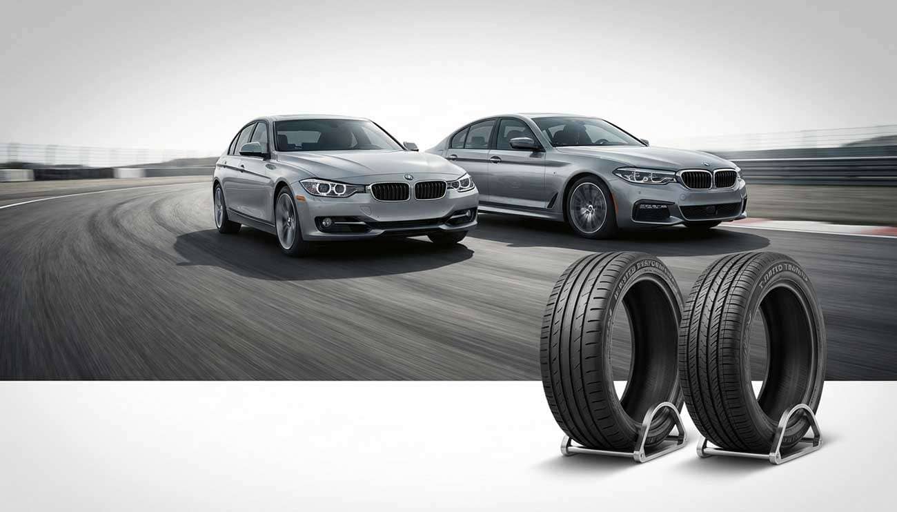 Two silver BMW cars driving side by side on a track with H and T rated tires in the foreground for comparison