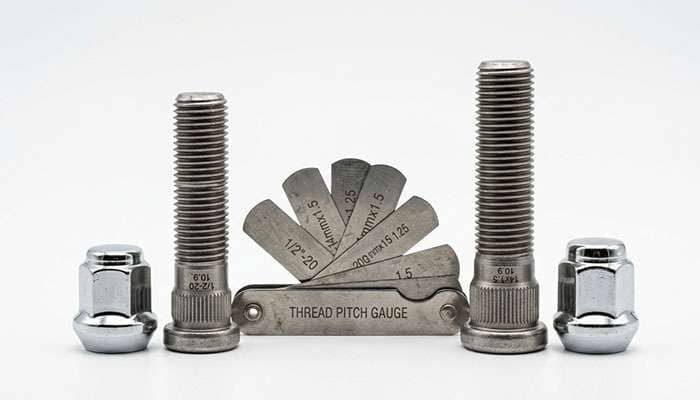 Half-inch and 14 mm Jeep wheel studs with matching cone-seat lug nuts side by side next to a thread pitch gauge