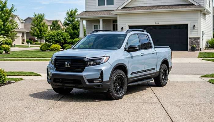 Light blue Honda Ridgeline pickup truck with off-road tires parked on a concrete driveway in a suburban neighborhood.