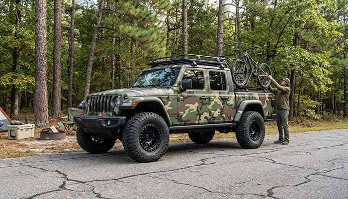 Camouflage Jeep truck with 37-inch tires and a mountain bike mounted on the roof parked on asphalt near trees