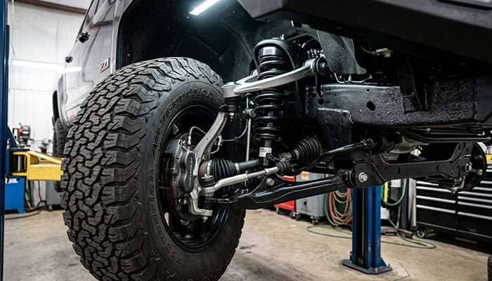Front suspension of a leveled pickup truck showing the CV axle, upper ball joint, and strut spacer behind a larger all-terrain tire