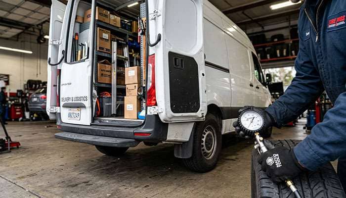 Technician checking rear tire pressure on a loaded high-roof cargo van with shelving visible through the open rear doors