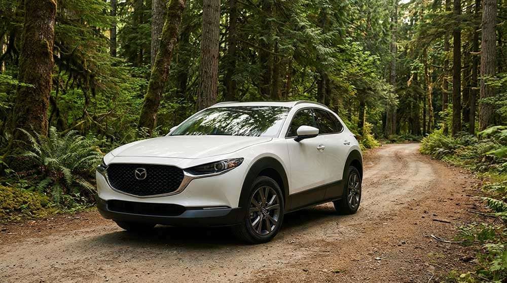 White 2022 Mazda CX-30 with 18 inch wheels parked on dirt road surrounded by dense green forest