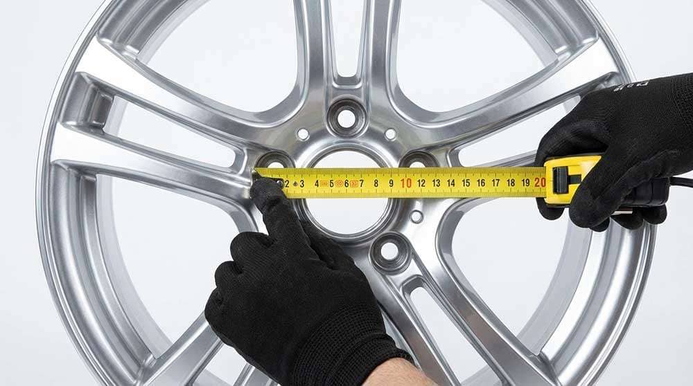 Measuring the wheel bolt pattern on a silver alloy wheel using a yellow tape measure for accurate fitment