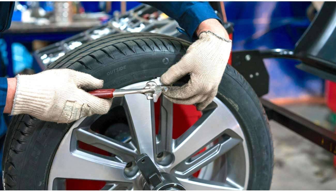 A mechanic wearing gloves installs a wheel weight on a car tire rim using pliers in a workshop