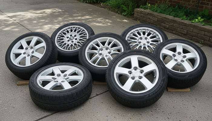 Seven silver OEM Mazda and Honda wheels some with tires arranged on grass in dim light