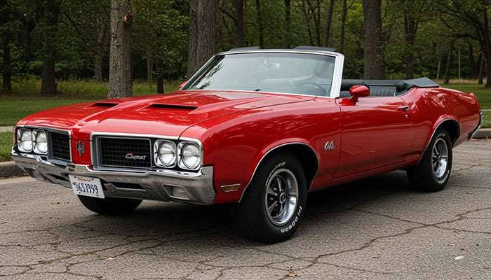 Red 1970 Oldsmobile 442 convertible muscle car with black racing stripes parked on a cracked pavement near trees