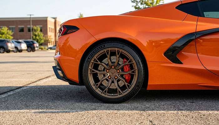 Close-up of the rear wheel and body of an orange Corvette C8 with custom bronze multi-spoke rims in a parking lot