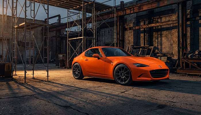 Orange Mazda Miata with custom deep-dish wheels parked in an industrial setting with scaffolding and shadows