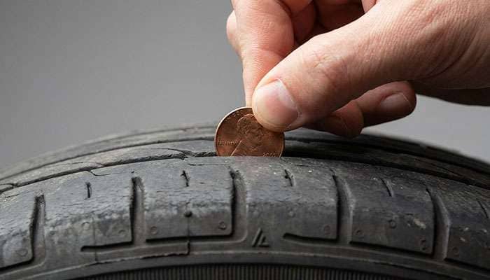 Hand holding a penny to check tire tread depth on a worn car tire at home.