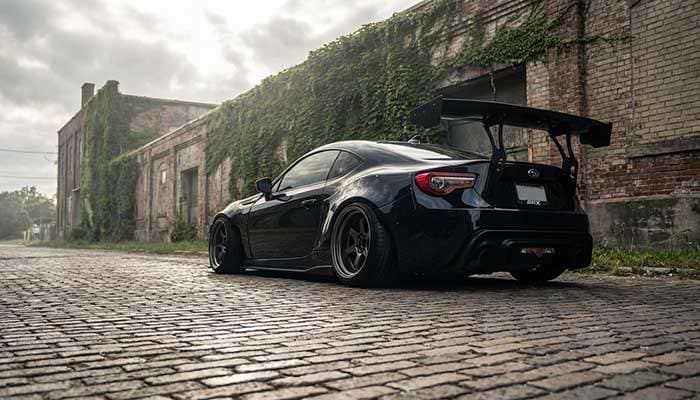 Rear view of lowered black sports car with wide wheels and large spoiler parked on brick road by building