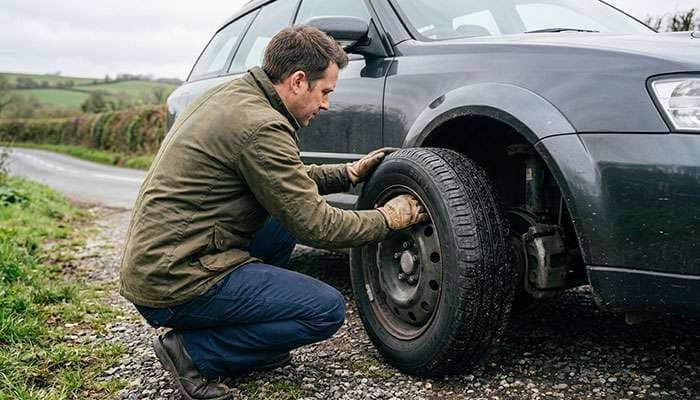 Person crouching beside a car inspecting the tire sidewall and wheel rim after a pothole impact