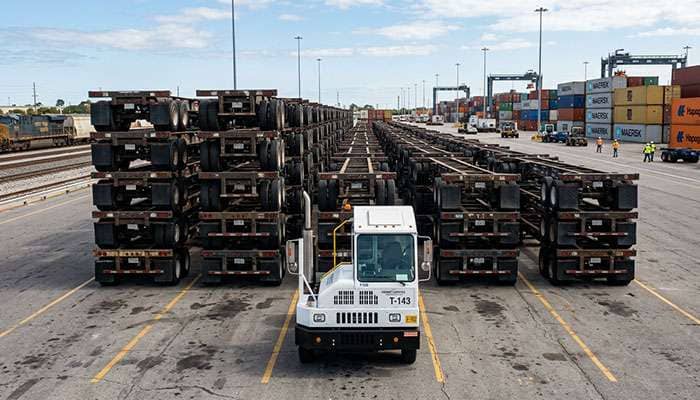 Rows of stacked shipping container chassis with a white rail terminal truck parked in front at a freight yard.