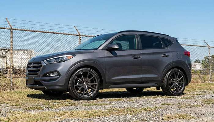 Silver Hyundai Tucson with aftermarket wheels parked on grassy gravel near a chain-link fence under clear sky