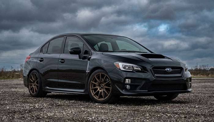 Black Subaru WRX sedan with aftermarket wheels parked on gravel under cloudy sky with wheel fitment guide text overlay