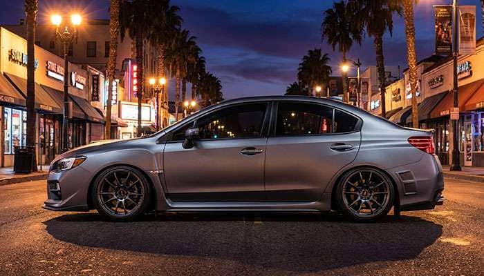 Side view of a modified gray Subaru WRX with aftermarket wheels parked at night near palm trees and a stop sign