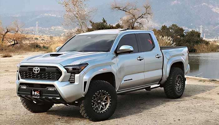 Silver Toyota Tacoma with Hardrock Offroad wheels parked on a dirt area near a lake and mountains in the background
