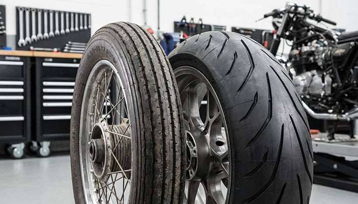 Close-up comparison of vintage motorcycle front tire next to modern wider tire showing size and tread pattern differences