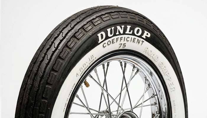 Close-up of a vintage motorcycle tire with a whitewall and period-style raised sidewall lettering against a clean white studio backdrop