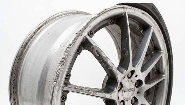 Close-up of an aluminum wheel showing visible curb rash damage and clear coat peeling on the lip and spokes against a clean white studio backdrop