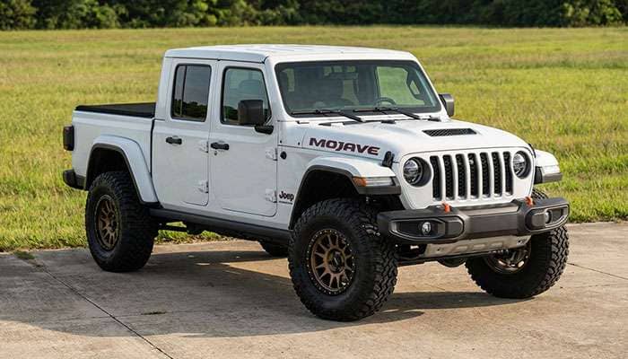 White Jeep Gladiator Mojave with large off-road tires and bronze wheels parked on concrete near a grassy area