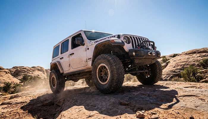 White Jeep Wrangler with large off-road tires parked on rocky desert terrain under clear blue sky