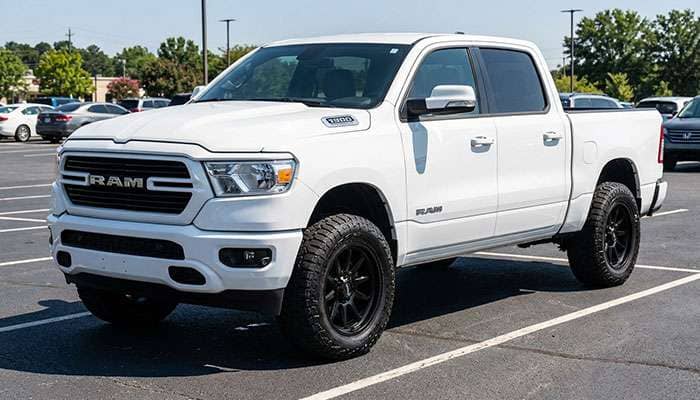 White 4th Gen Ram 1500 truck with black aftermarket wheels and all-terrain tires parked in a lot on a sunny day