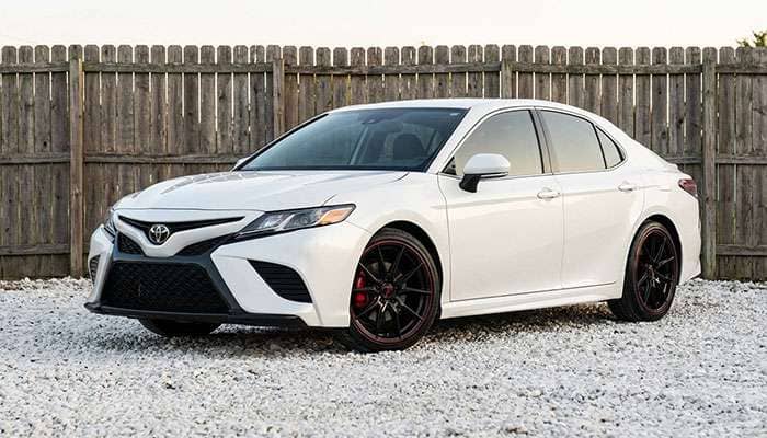 White Toyota Camry with black and red aftermarket rims parked on white gravel in front of a wooden fence