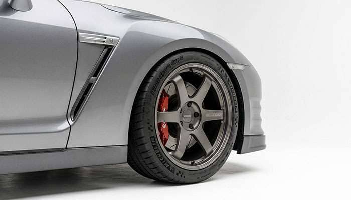 Front three-quarter view of a wider aftermarket tire fitted under a vehicle fender showing tight clearance and aggressive stance on a white studio backdrop