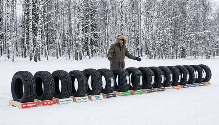 Person presenting a lineup of 11 winter tires outdoors in snowy landscape with birch trees in the background