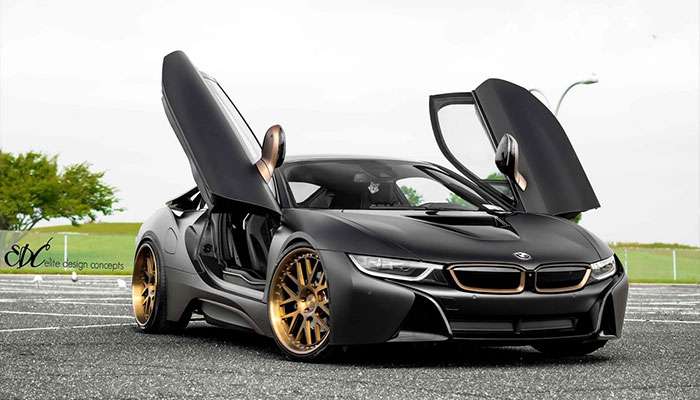 Matte black BMW i8 with bronze wheels and butterfly doors open parked on asphalt with greenery in the background