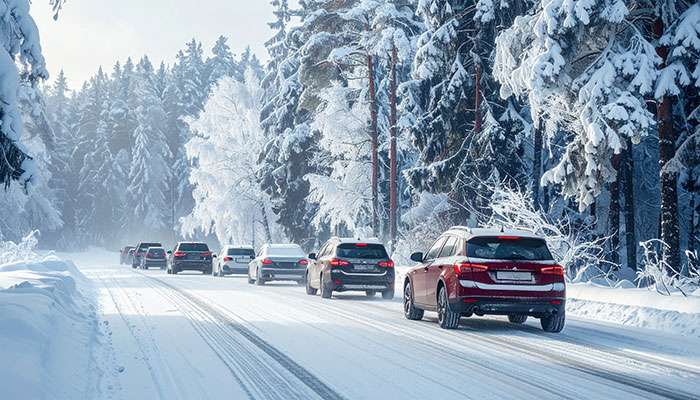 Line of cars driving cautiously on a snowy road surrounded by snow-covered trees in winter conditions