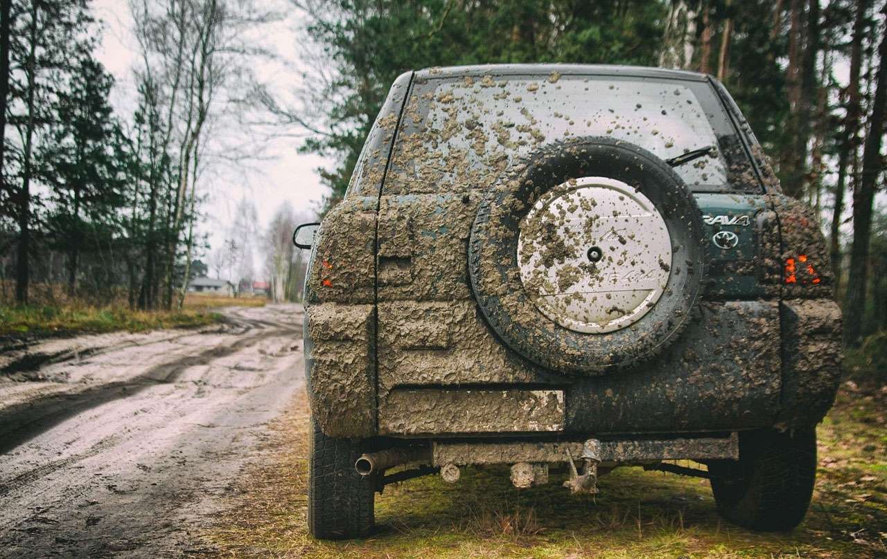 Rear view of a mud-covered Toyota RAV4 4x4 parked on a dirt road in a forested area.