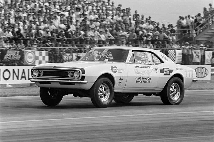 Classic drag racing car with sponsor decals speeding on track in front of a large crowd at a race event