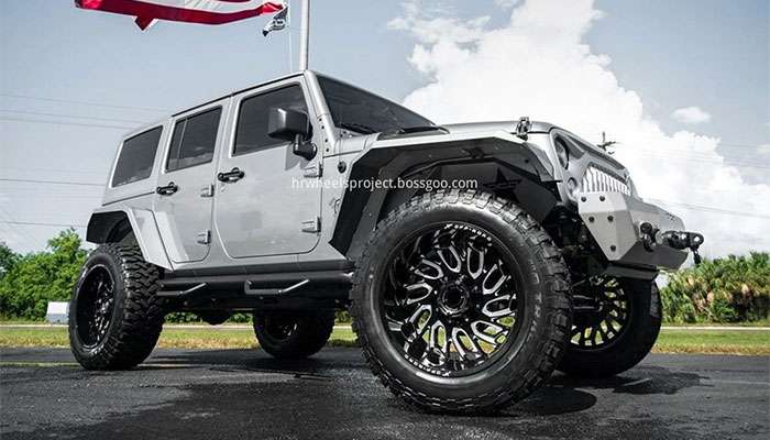 Silver Jeep Wrangler with large off-road tires and an American flag in the background on a sunny day.