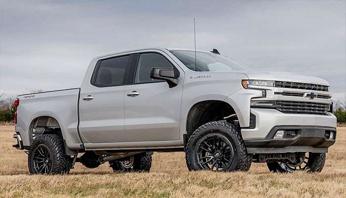 Silver Chevrolet Silverado pickup truck with black Fuel Off-Road wheels parked on dry grass under a cloudy sky.